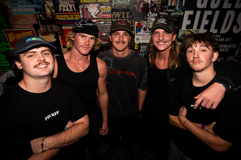 Close-up portrait of five members of indie band Fin Key standing together in front of a wall covered in gig posters.